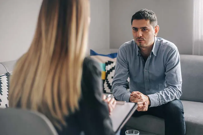 A man sits on a couch talking to a woman with a notepad, suggesting a counseling or therapy session in a living room setting.