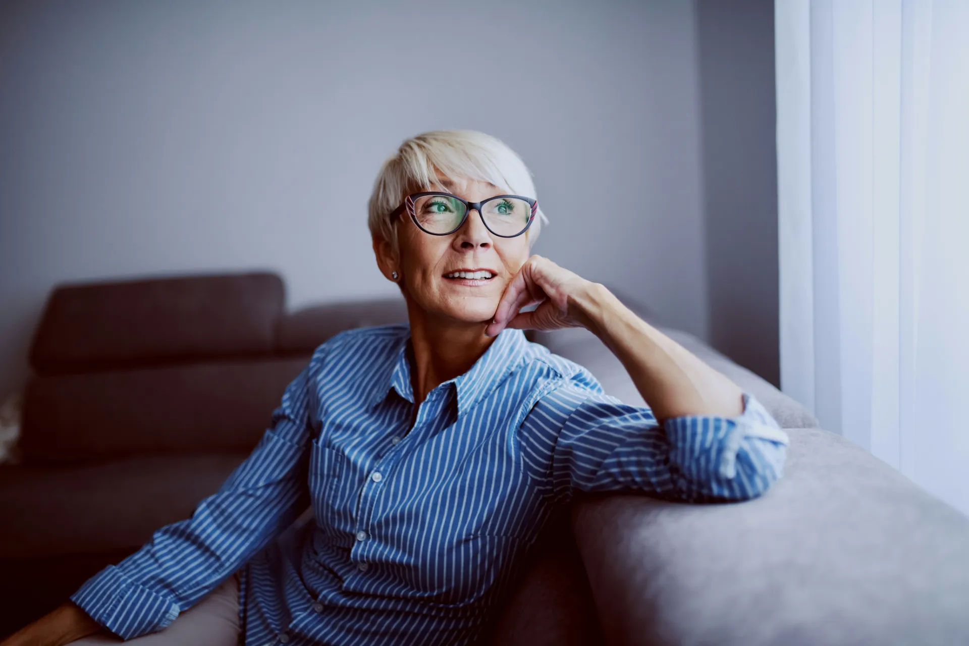 Portrait of a smiling senior woman with glasses, sitting on a sofa in a living room and looking out a nearby window.
